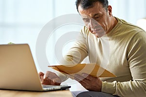 Portrait of busy latin middle aged businessman looking at the envelope, using laptop while working from home