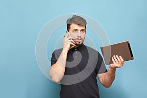 Portrait of a busy man standing on a blue background with a tablet in his hands and talking on the phone.Business man with gadgets