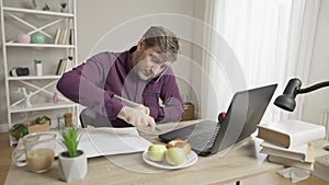 Portrait of busy man talking on the phone, using laptop and tablet, and writing down notes. Overworking Caucasian