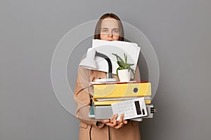 Portrait of busy young adult business woman holding moving box with stuff at new office and papers in her mouth, looking at camera