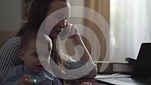 Portrait of busy young mother working at home with the baby on her laps. The woman talking by cellphone while her son