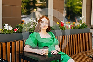 Portrait of calm attractive young woman holding cup with tasty coffee sitting at table in outdoor cafe terrace in summer
