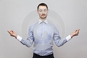 Portrait of calm handsome bristle businessman in classic light blue shirt standing in yoga pose with closed eyes and meditating