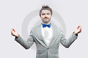 Portrait of calm relax handsome bearded man in casual grey suit and blue bow tie standing with raised arms, closed eyes and doing
