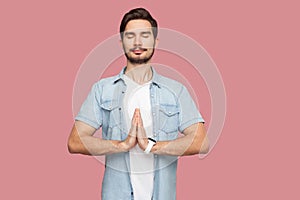 Portrait of calm relax handsome bearded young man in blue casual style shirt standing with palm hands, closed eyes and doing yoga