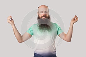 Portrait of calm relaxed middle aged bald man with long beard in light green t-shirt standing with closed eyes, raised arms and