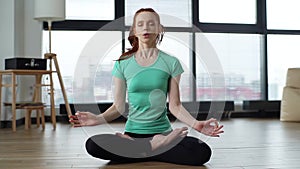 Portrait of calm relaxed young woman meditating in lotus position sitting on floor with closed eyes at home practising