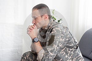Portrait of calm serious Caucasian military man wearing camouflage uniform and cap sitting on a sofa, having depressed