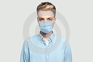 Portrait of calm serious handsome young man with surgical medical mask in light blue shirt standing and looking at camera with