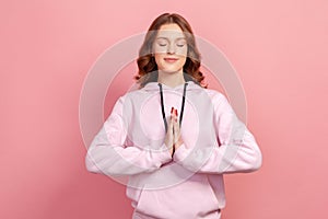 Portrait of calm teen girl in hoodie concentrating her mind, keeping hands namaste gesture, meditating, yoga exercise breath