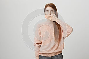 Portrait of calm tired ginger girl covering mouth with hand, standing over gray background in casual outfit. Body
