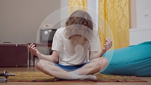 Portrait of calm young retro man with long curly hair falling asleep sitting in lotus pose indoors. 1990s guy practicing