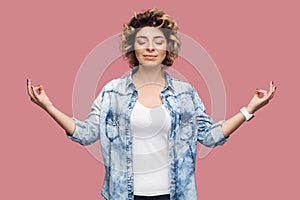 Portrait of calm young woman with curly hairstyle in casual blue shirt standing with closed eyes, raised arms, smile and doing