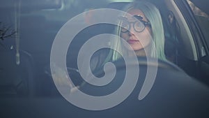 Portrait through car window of young cheerful blonde driver girl with smartphone in hands, wearing sunglasses and jeans jacket.