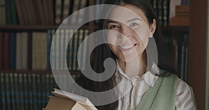 Portrait of carefree Asian girl student standing in university library holding books smiling looking at camera