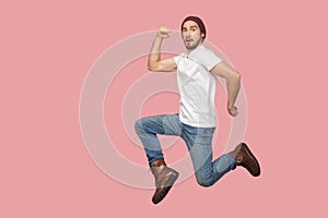 Portrait of carefree bearded hipster young man in white shirt and blue jeans with hat jumping in super mario pose and looking at