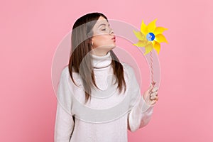 Portrait of carefree brunette woman blowing at paper windmill, playing with pinwheel toy on stick.