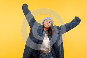 Portrait of carefree cheerful girl wearing warm winter hat and fur coat raising hands up, smiling joyfully