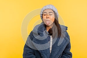 Portrait of carefree girl wearing warm winter hat and fur coat, stylish urban outfit, standing with closed eyes showing tongue