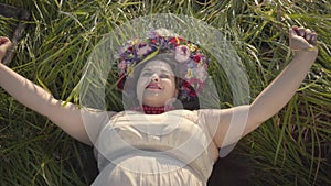 Portrait of carefree overweight woman with a wreath on her head lying on the grass in summer field holding the scythe