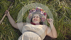 Portrait of carefree overweight woman with a wreath on her head lying on the grass in summer field holding the scythe