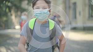 Portrait of carefree schoolboy in face mask running along schoolyard in sunlight with blurred classmates at the