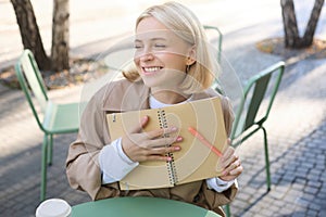 Portrait of carefree, smiling young woman with notebook and pen, sitting in outdoor cafe near table, writing in journal