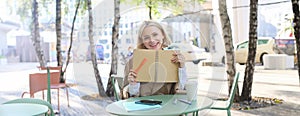 Portrait of carefree, smiling young woman with notebook and pen, sitting in outdoor cafe near table, writing in journal