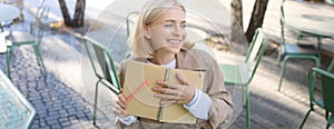 Portrait of carefree, smiling young woman with notebook and pen, sitting in outdoor cafe near table, writing in journal