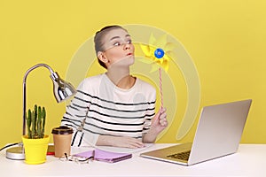 Woman blowing at windmill while sitting on workplace with laptop, playing with pinwheel toy on stick