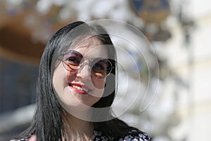 Portrait of carefree young woman smiling and looking at camera with urban background. Cheerful european girl wearing sunglasses in