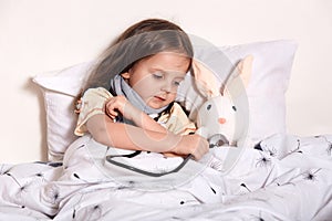 Portrait of caring sweet kid with scarf around neck lying in her bed with favourite toy, examining it with stethoscope, looking