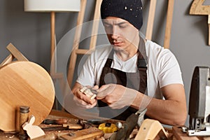 Portrait of carpenter man in brown apron making a handmade wooden toy in a home workshop, handyman carves a toy in wood with a