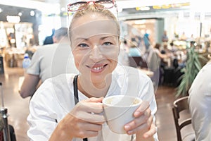 Portrait of a casual young blond woman having a cup of coffee, sitting in cafe indoors of an airport, station, food