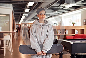 Portrait Of Casually Dressed Young Businesswoman With Skateboard Standing In Open Plan Workplace