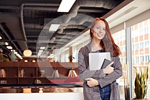 Portrait Of Casually Dressed Young Businesswoman Standing In Modern Open Plan Workplace