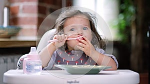 portrait caucasian baby girl about 2 years old in bib eating pasta from plate sitting high chair, self feeding for kids
