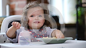 portrait caucasian baby girl about 2 years old in bib eating pasta from plate sitting high chair, self feeding for kids