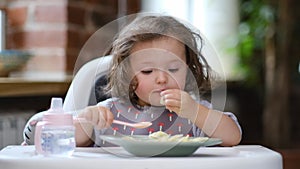 portrait caucasian baby girl about 2 years old in bib eating pasta from plate sitting high chair, self feeding for kids