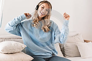 Portrait of caucasian blond girl 20s wearing headphones smiling and listening to music at home