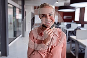Portrait of caucasian businesswoman wearing lowered face mask standing in office smiling to camera