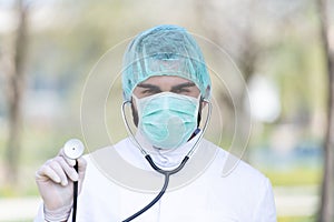 Portrait of a Caucasian Doctor male with mask, scrub cap holding a stethoscope in the park
