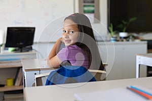 Portrait of caucasian elementary schoolgirl looking over shoulder while sitting at desk in class