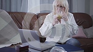 Portrait of Caucasian fenmale retiree in eyeglasses sitting at couch with coffee cup and using laptop. Blond Caucasian