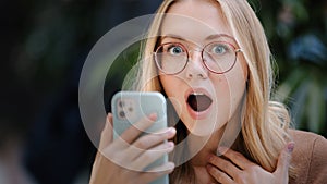 Portrait caucasian girl blonde successful lucky woman in glasses looks at screen of mobile phone wins online message