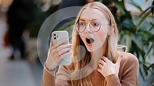 Portrait caucasian girl blonde successful lucky woman in glasses looks at screen of mobile phone wins online message