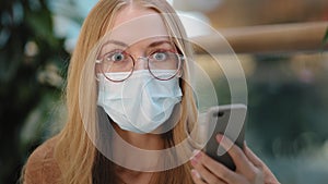 Portrait caucasian girl in glasses wears face protective medical mask looks at screen of mobile phone woman feels shock