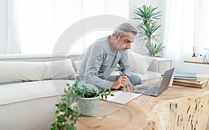Portrait of Caucasian grey hair man writing notes in notepad while using laptop from workspace in living room at home interior.