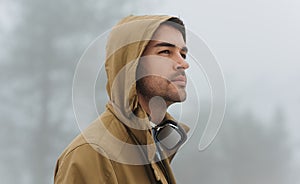 Portrait of Caucasian handsome male walking in misty nature, listening to music on his headphones outside. Traveler man wearing