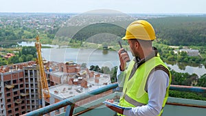 Portrait of Caucasian handsome man builder manager in uniform and helmet looking at digital tablet and talking in walkie